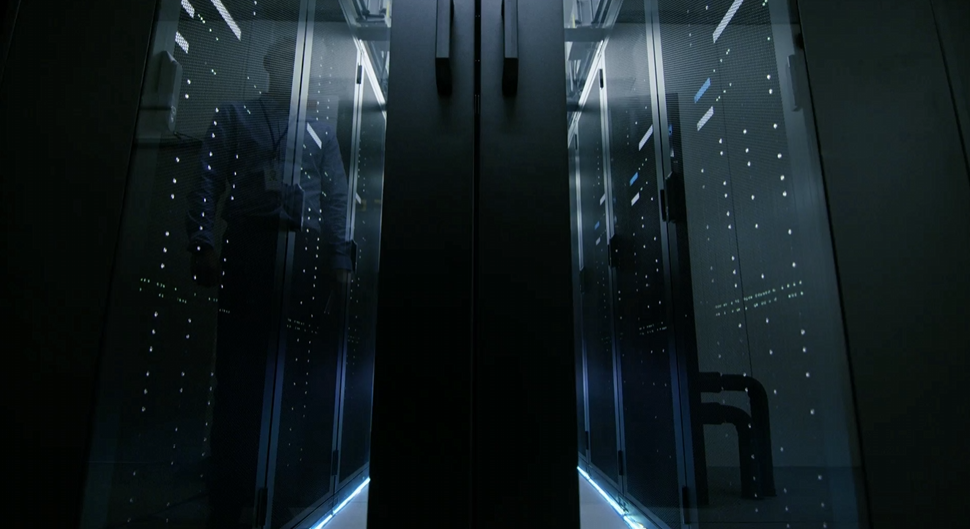 Darkened aisle between rows of server racks in a modern data centre, illuminated by cool blue lighting.