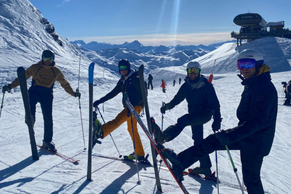 4 skiers posing with their skis on the mountain, the snow is white and fresh with a clear blue sky and bright sunshine
